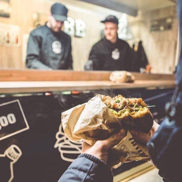 Eine Person hält einen halb gegessenen Burger in der Hand mit einer Serviette, im Hintergrund sind unscharf zwei Mitarbeiter im Inneren des Burgerbox Foodtrucks sichtbar.