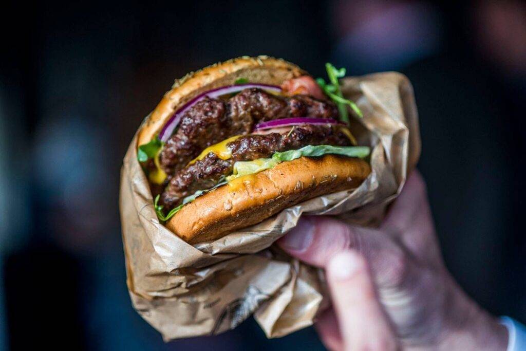 Hand hält einen appetitlichen Burger mit glänzendem Sesambrötchen, saftigem Rindfleischpatty, geschmolzenem Käse, frischem Salat und roten Zwiebelringen, eingewickelt in braunes Papier, im Fokus vor unscharfem Hintergrund.