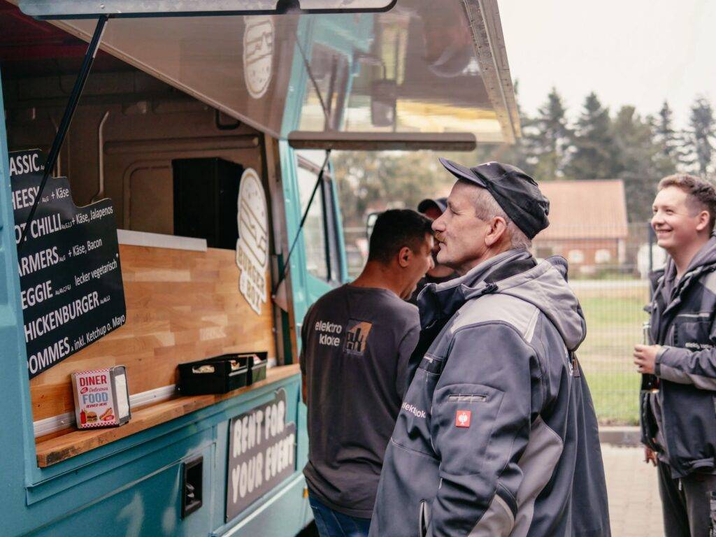 Kunden in Arbeitskleidung bestellen Essen am türkisfarbenen Burgerbox Foodtruck, der ein Menü mit verschiedenen Burgeroptionen anzeigt, während ein lächelnder junger Mann im Hintergrund wartet.