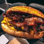 Nahaufnahme eines Gourmet-Burgers mit goldener Sesambrötchen, Schichten von geschmolzenem Käse, knusprigen Speckstreifen und saftigem Rindfleisch-Patty, serviert von Burgerbox.
