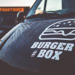 Vorderseite eines Foodtrucks mit dem Logo von Burgerbox, erkennbar an einem stilisierten Burger-Symbol und dem Schriftzug #BURGERBOX, im Hintergrund unscharf ein weiterer Foodtruck mit leuchtendem Schriftzug.
