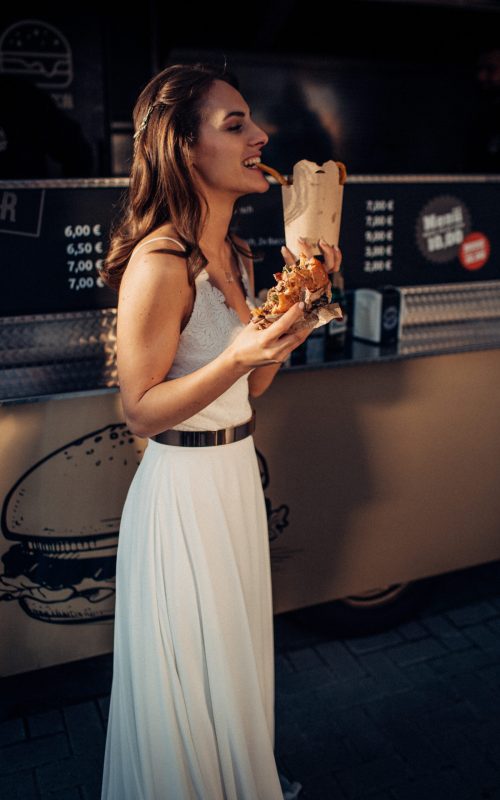 "Braut in weißem Kleid genießt Gourmet-Burger vor einem Foodtruck bei einer Outdoor-Catering-Veranstaltung in Osnabrück