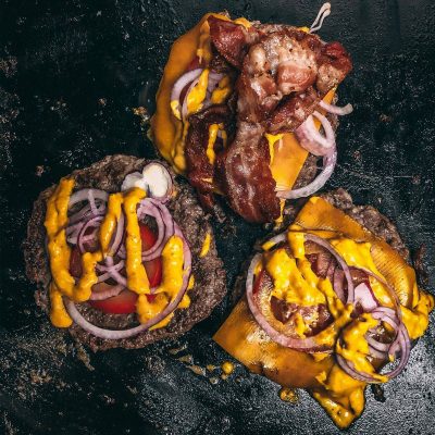 Drei saftige Cheeseburger-Patties auf einem Grill, garniert mit geschmolzenem Cheddar-Käse, roten Zwiebelringen und Senfsauce, in der Mitte mit knusprigem Bacon belegt, alles auf einer schwarzen Oberfläche mit Spritzern von Fett und Sauce.