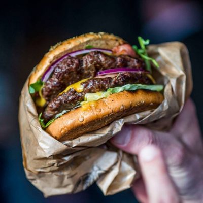 Hand hält einen appetitlichen Burger mit glänzendem Sesambrötchen, saftigem Rindfleischpatty, geschmolzenem Käse, frischem Salat und roten Zwiebelringen, eingewickelt in braunes Papier, im Fokus vor unscharfem Hintergrund.