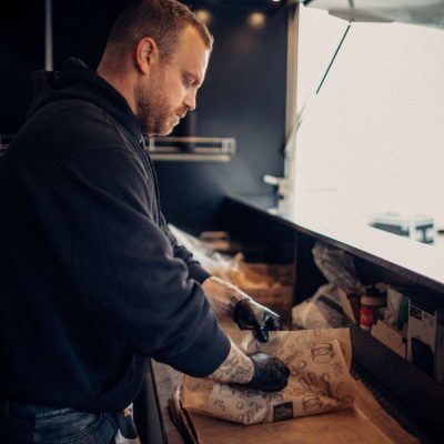 Ein Mann in Arbeitskleidung verpackt sorgfältig Lebensmittel in Papier in einem Foodtruck, wobei der Fokus auf den Händen und der Tätigkeit liegt, im Hintergrund ist die helle Öffnung des Verkaufsfensters zu sehen.