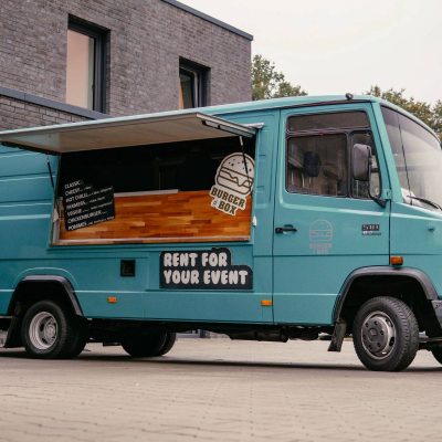 Türkisfarbener Burgerbox Foodtruck mit geöffnetem Verkaufsfenster und sichtbarem Menü, geparkt vor einem modernen Gebäude, mit dem Schriftzug "RENT FOR YOUR EVENT" an der Seite, bereit für Catering bei Veranstaltungen