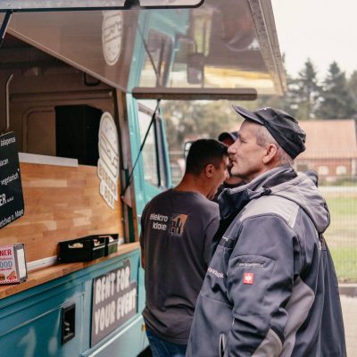 Kunden in Arbeitskleidung bestellen Essen am türkisfarbenen Burgerbox Foodtruck, der ein Menü mit verschiedenen Burgeroptionen anzeigt, während ein lächelnder junger Mann im Hintergrund wartet.
