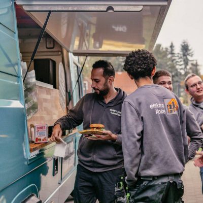 Ein Mann in Arbeitskleidung erhält einen Burger von einem Mitarbeiter im Inneren eines türkisfarbenen Burgerbox Foodtrucks, während andere Kunden warten und im Hintergrund zu sehen sind.